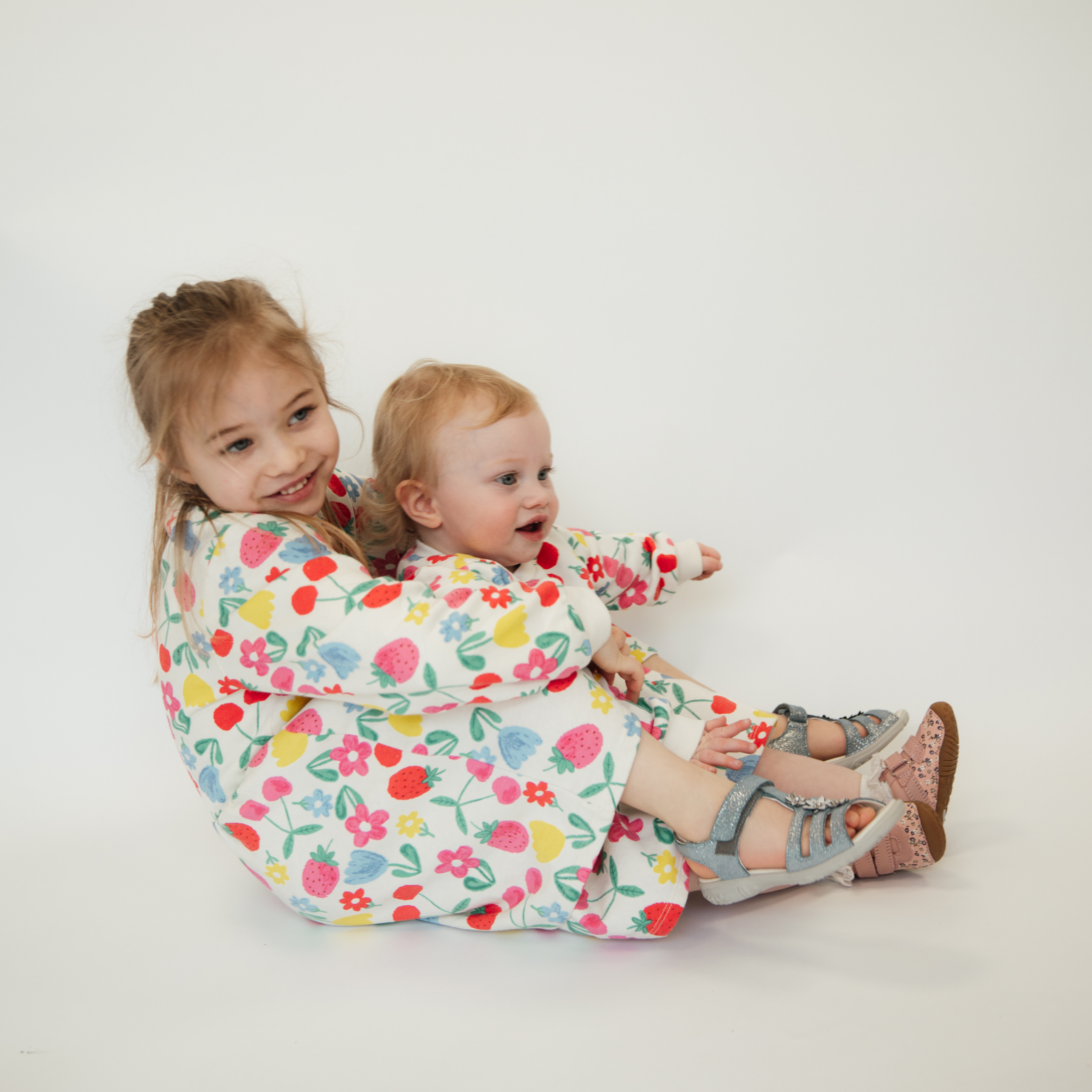 Two young sisters sitting together wearing matching floral dresses, the toddler wearing silver sandals.