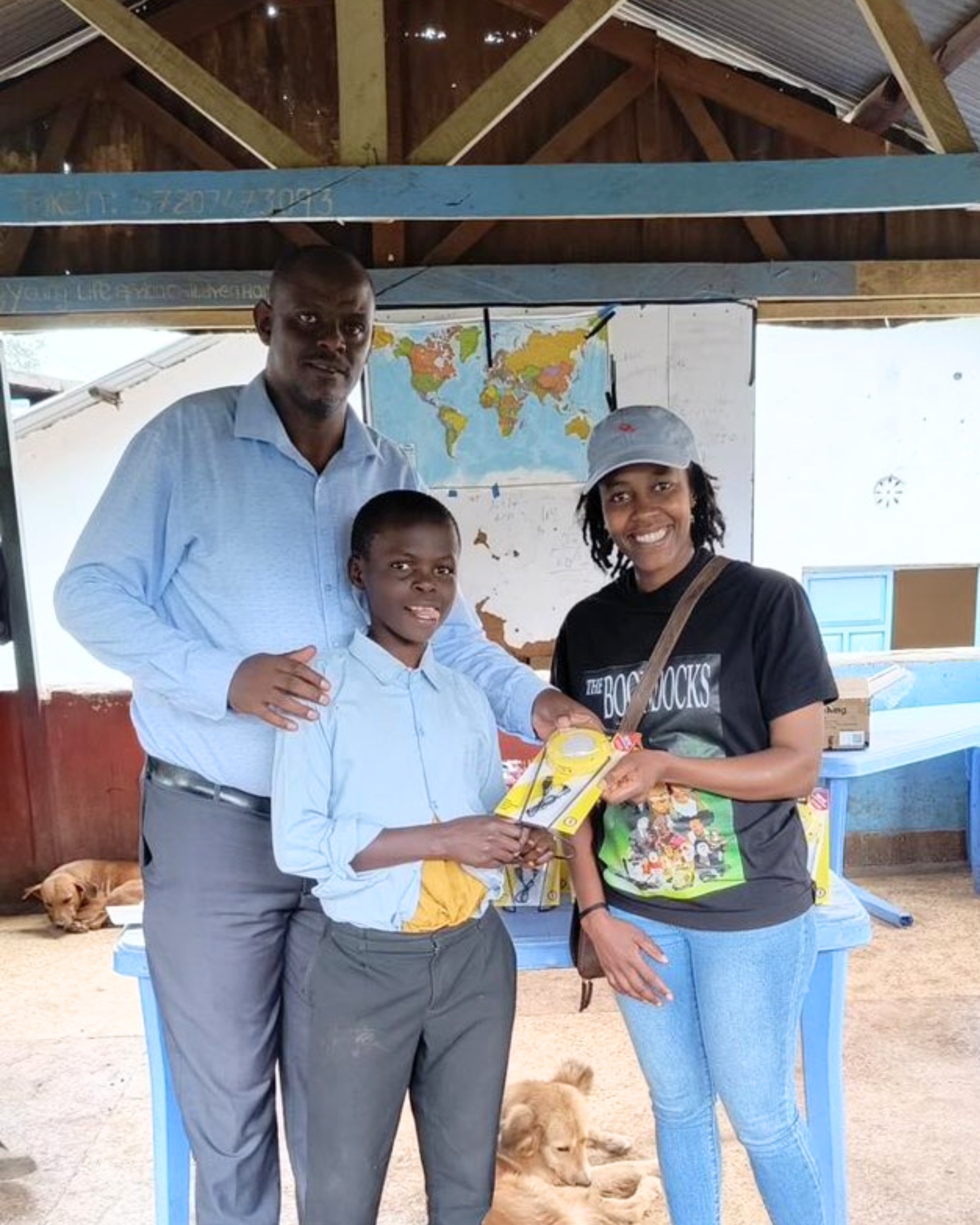 Three people posing together indoors, with a child receiving yellow books from a smiling woman in a cap and black t-shirt.