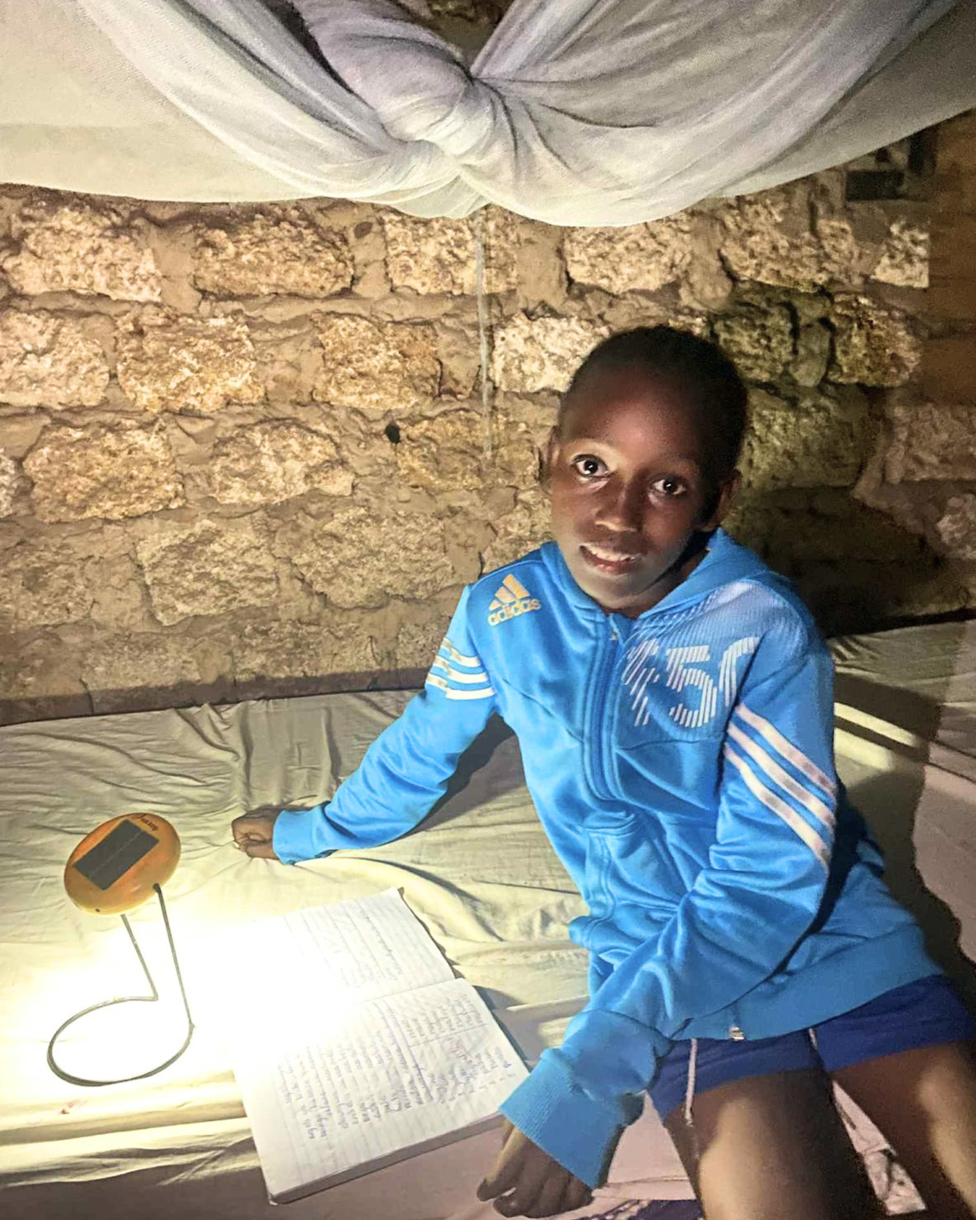 Child studying at night by solar lamp light, sitting on bed with open notebook in stone-walled room