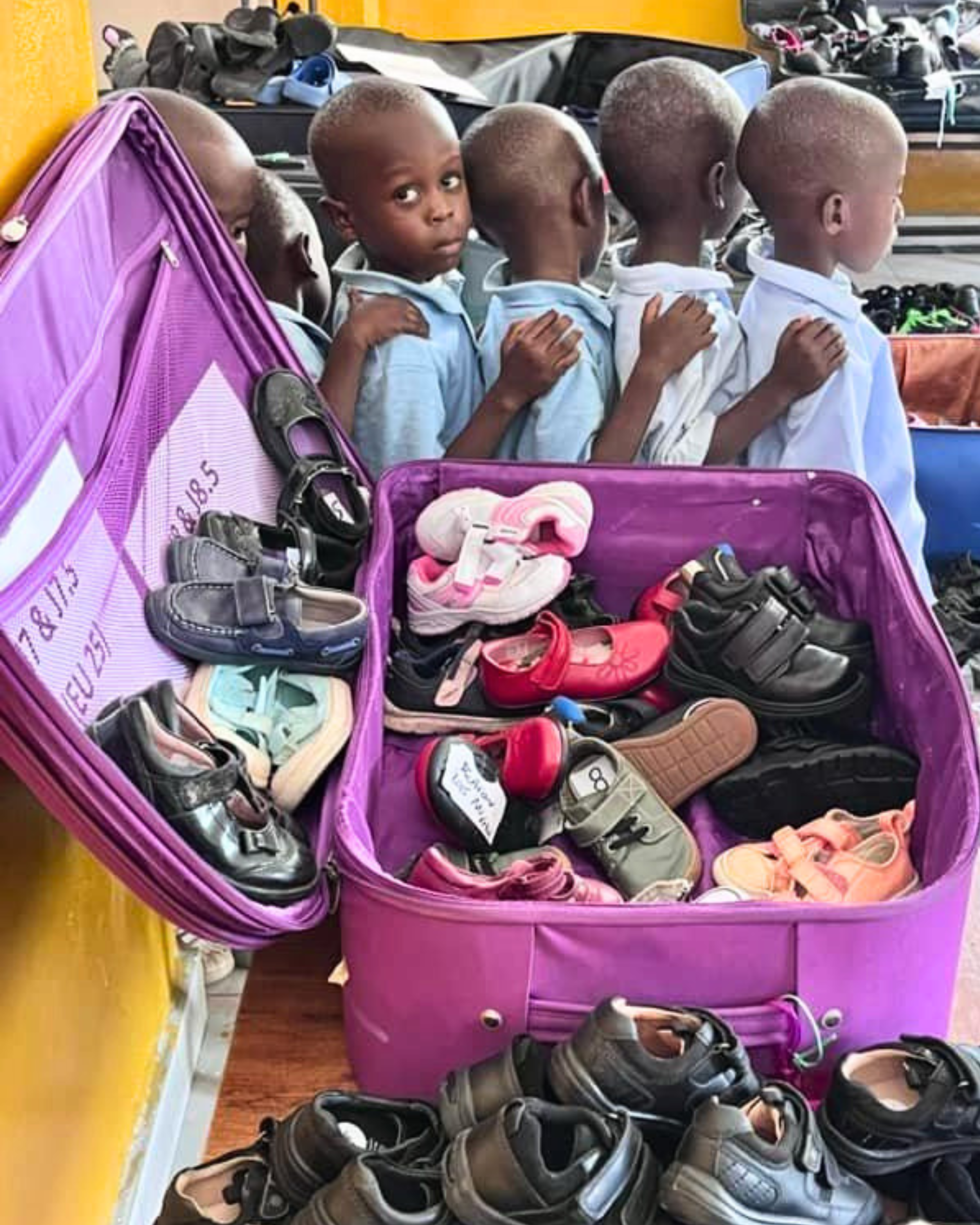 Children in blue uniforms selecting shoes from an open purple suitcase filled with donated footwear.