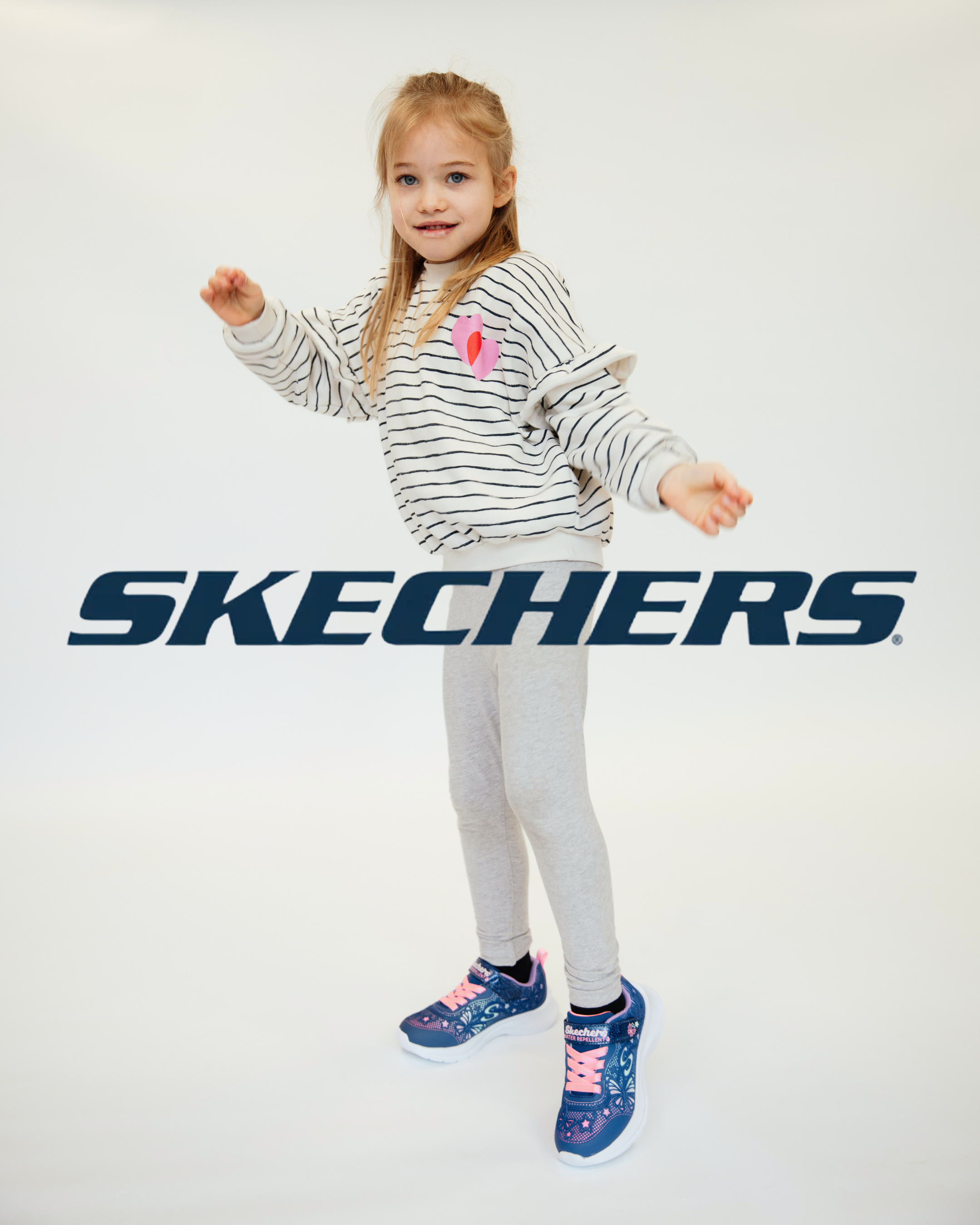 Girl wearing navy/pink Skechers trainers with butterfly print, grey leggings and striped sweatshirt, posed playfully