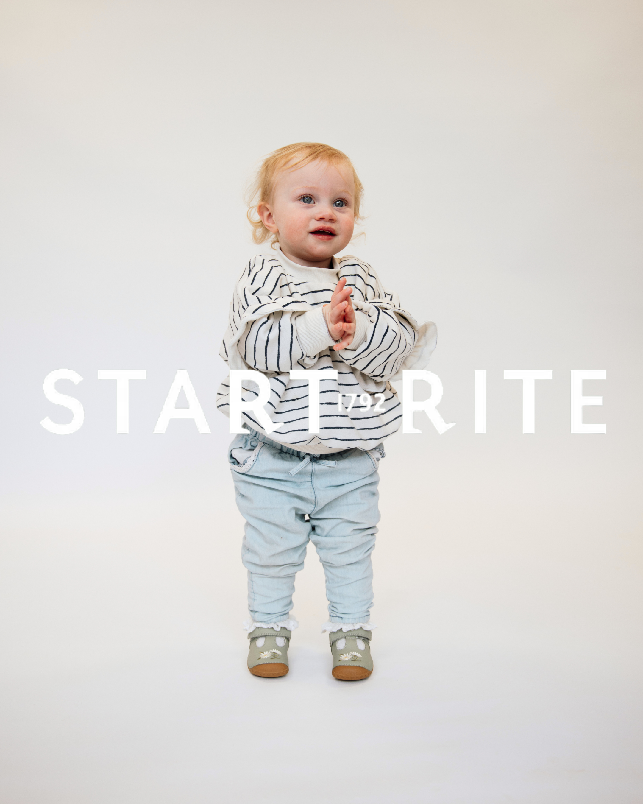 Toddler standing and clapping wearing striped sweatshirt, light jeans and sage green Start-Rite first shoes