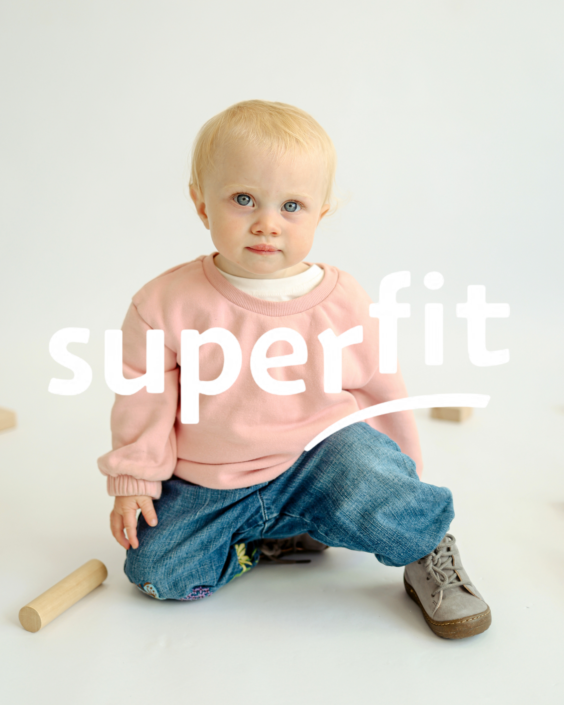 Toddler with blonde hair sitting on floor wearing a pink sweatshirt, jeans and grey Superfit boots.