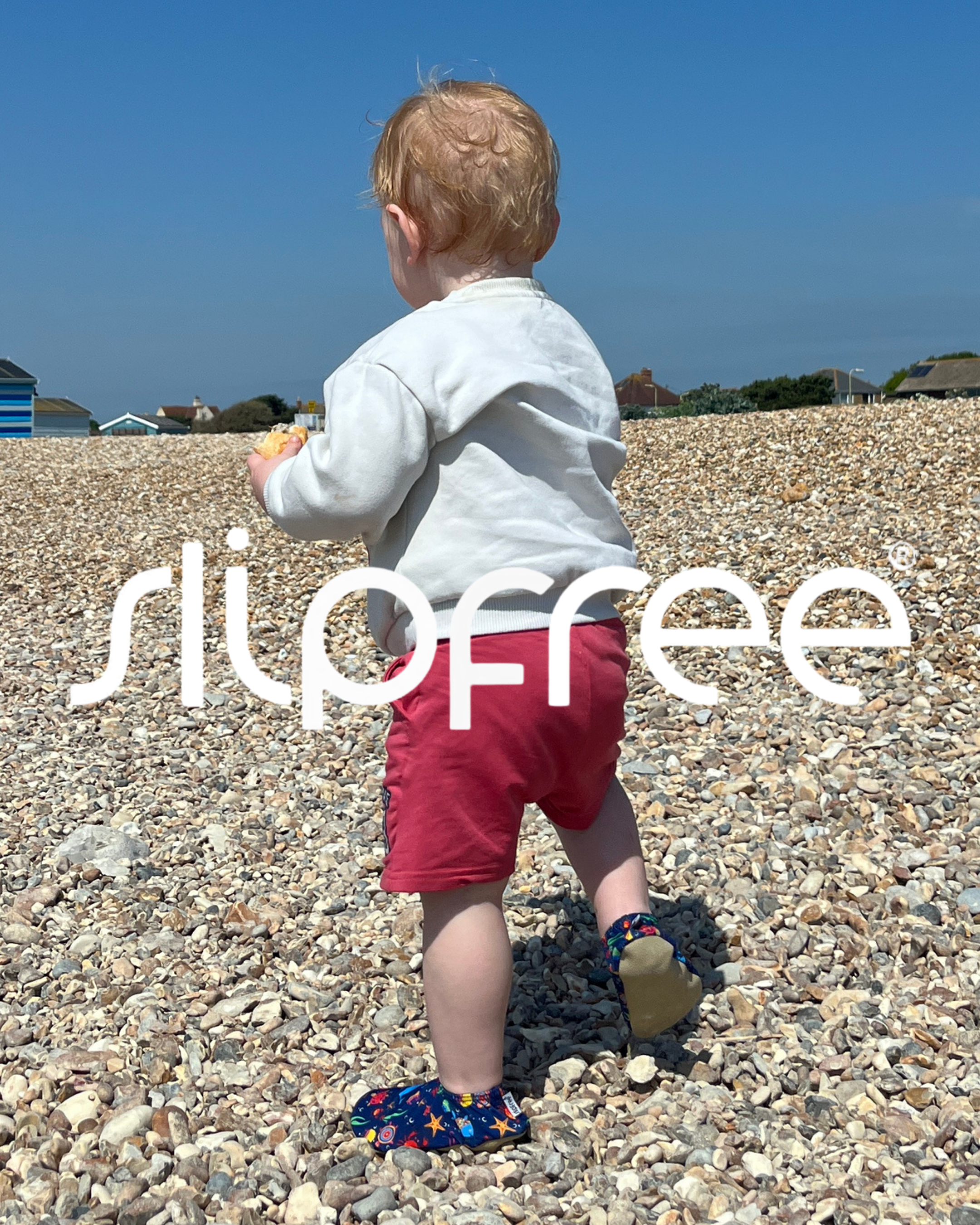 Toddler walking on a pebble beach wearing blue Slipfree shoes, white top and red shorts on a sunny day.