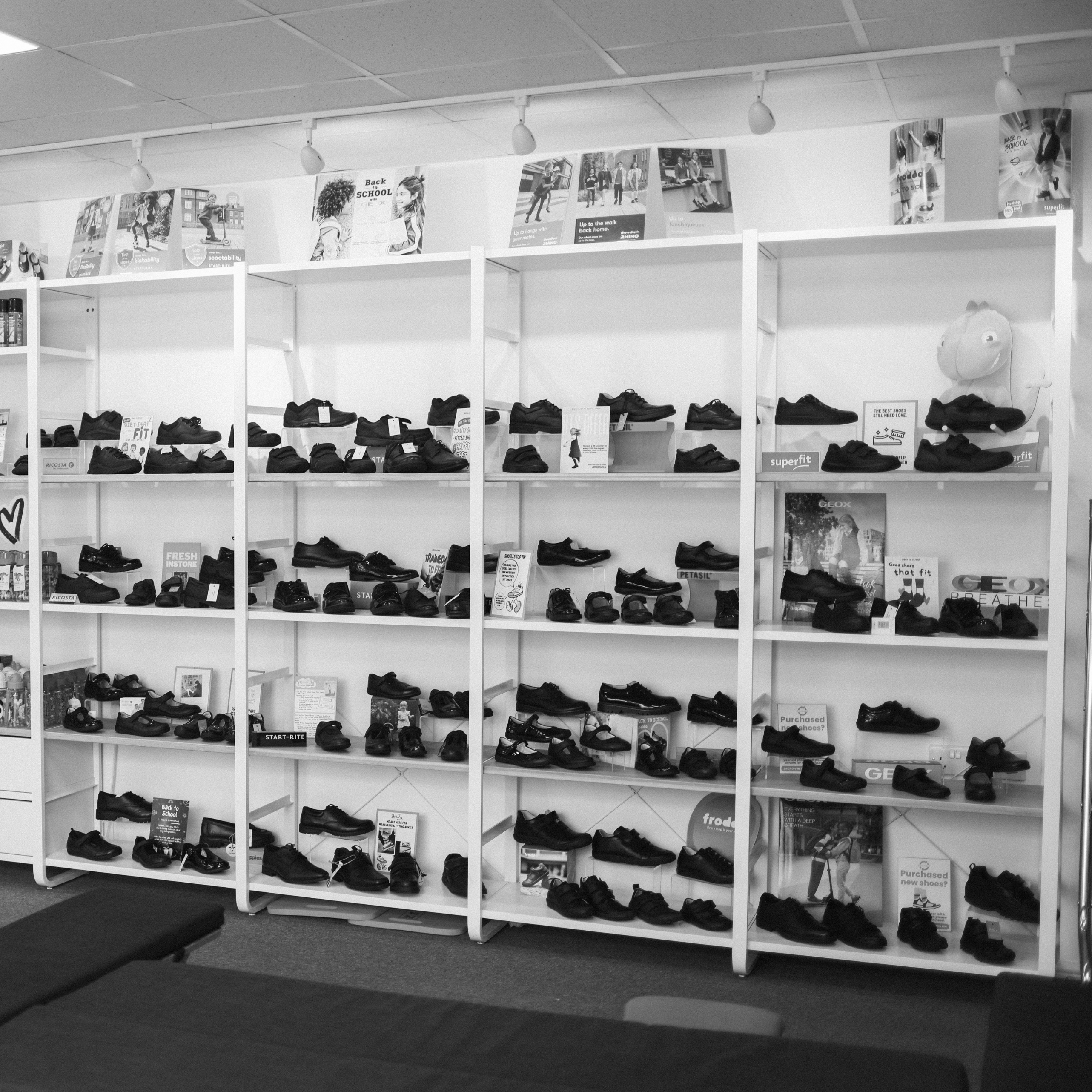 White shelving units displaying rows of children's black school shoes in a ShuZu store, with Back to School signage.