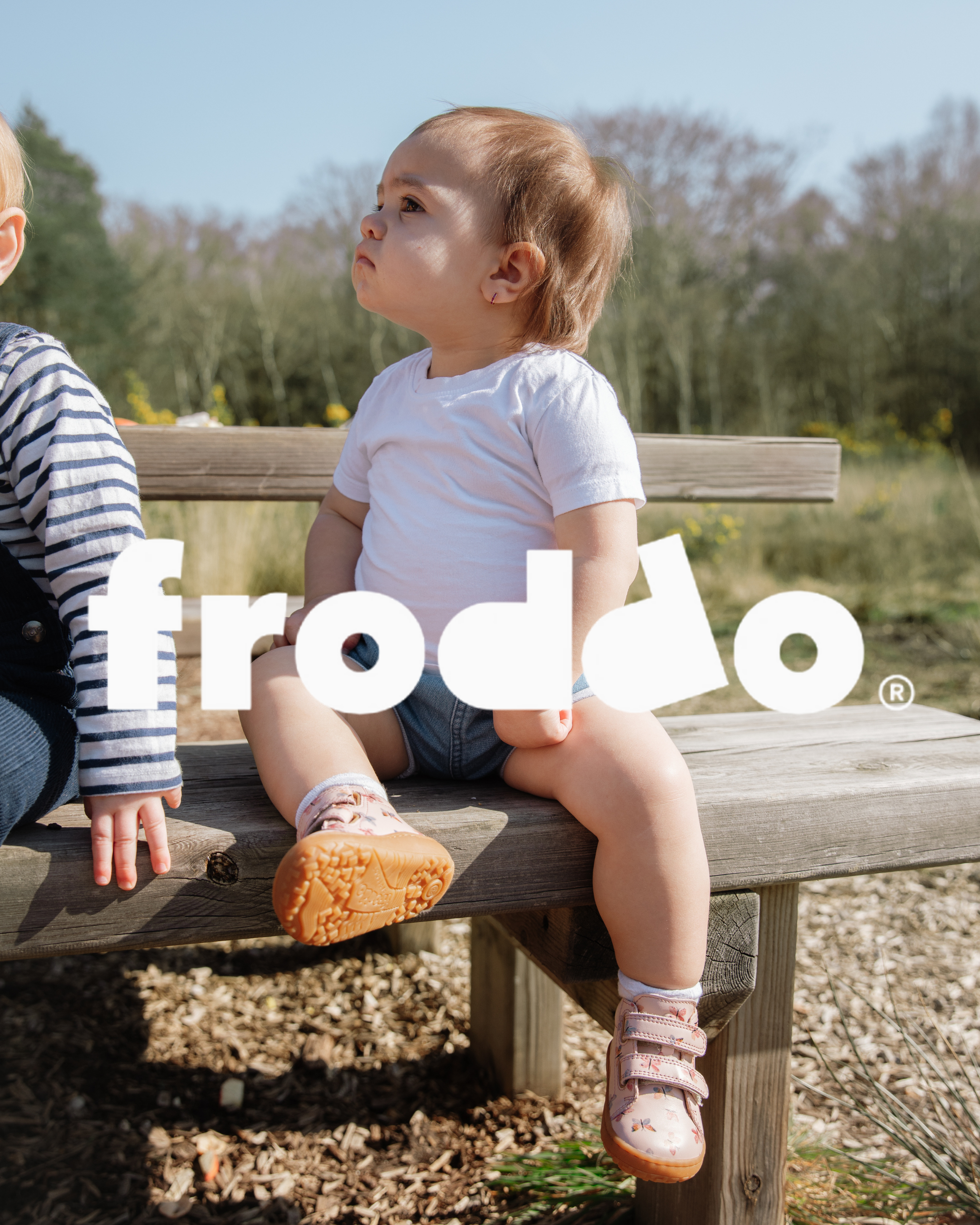 Toddler sitting on a wooden bench outdoors wearing Froddo pink sneakers with velcro straps and orange soles.