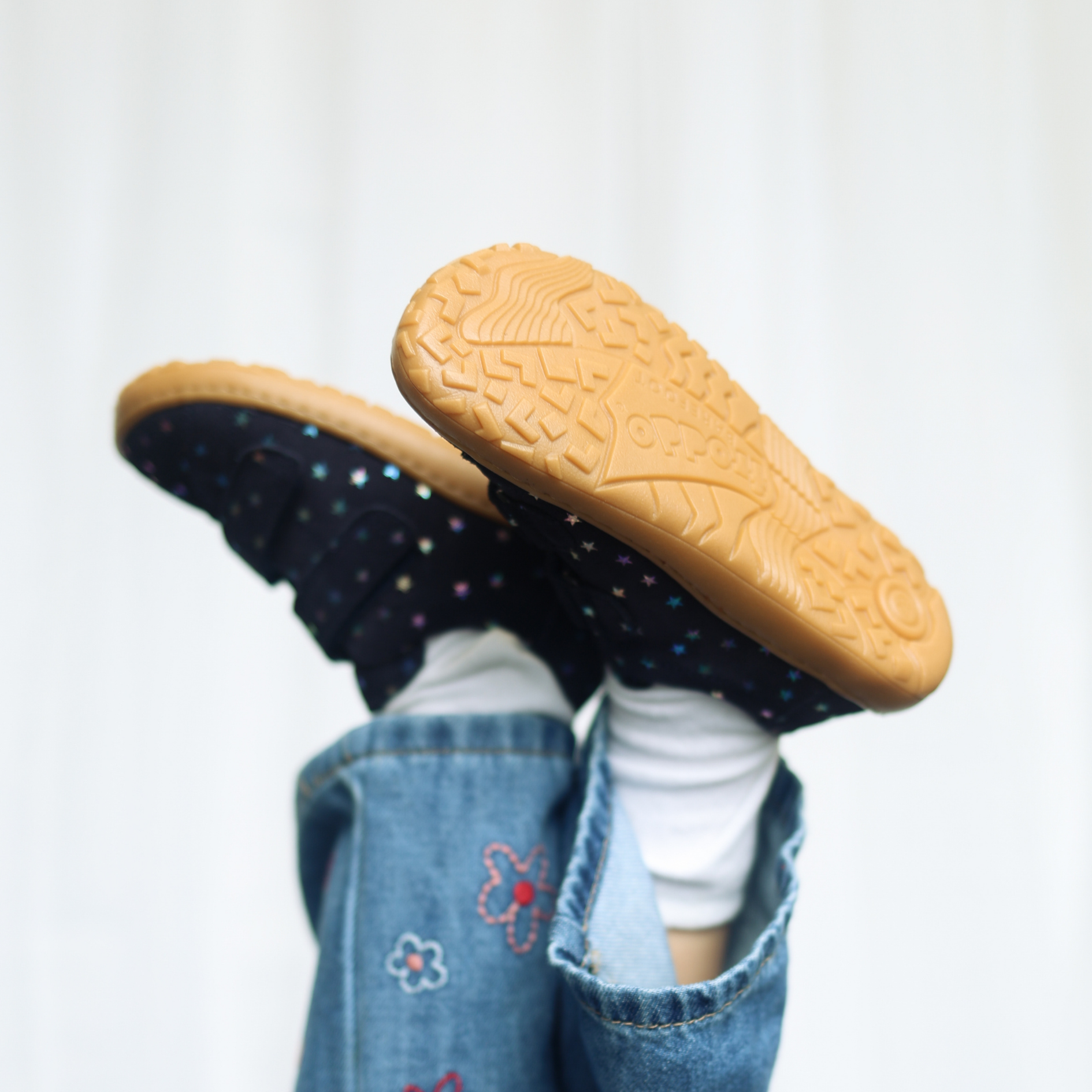 Child's feet raised up showing soles of navy star-print shoes with tan rubber grip soles, wearing floral denim dungarees.