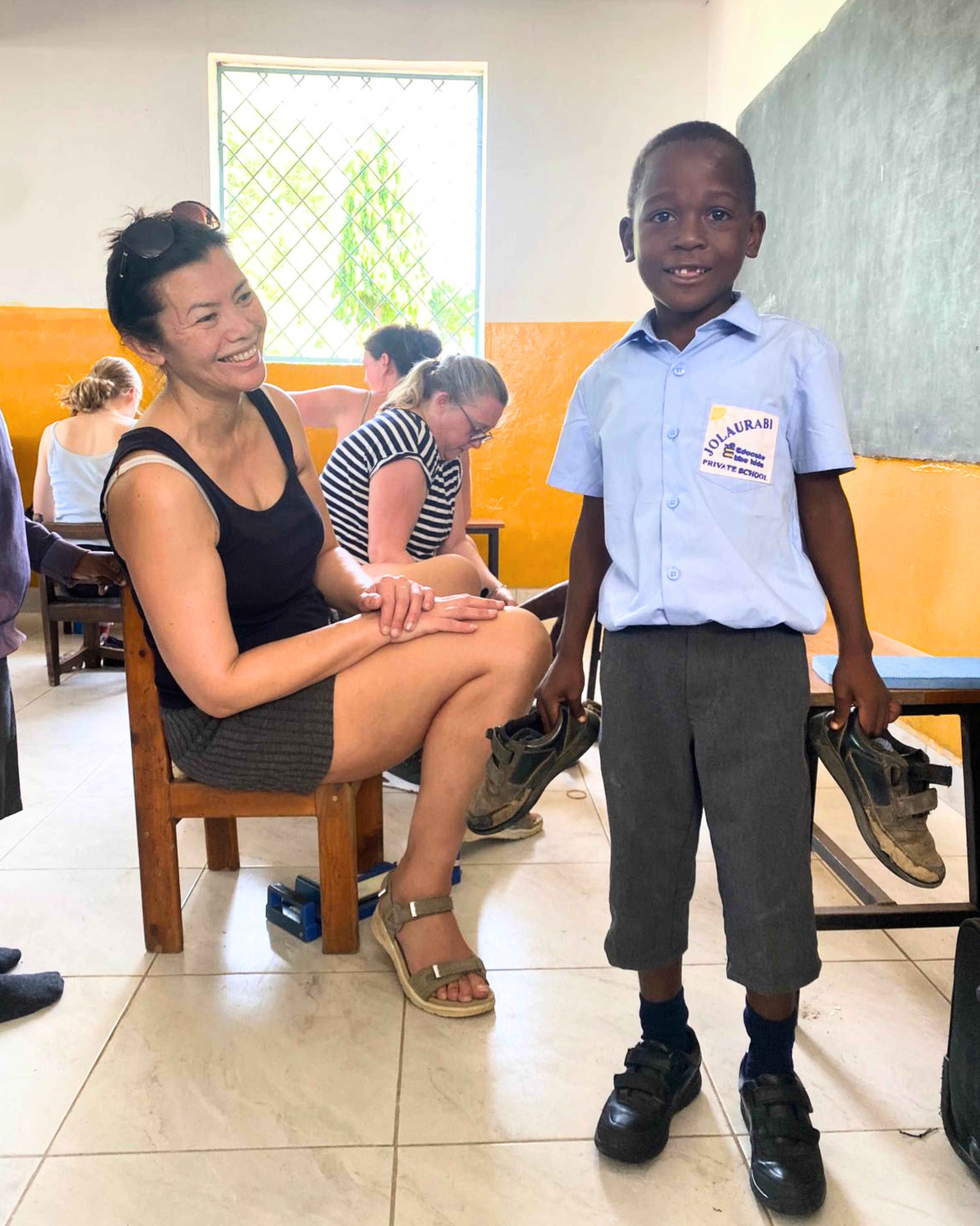 A woman smiles beside a young boy in school uniform proudly holding new black shoes in a classroom.