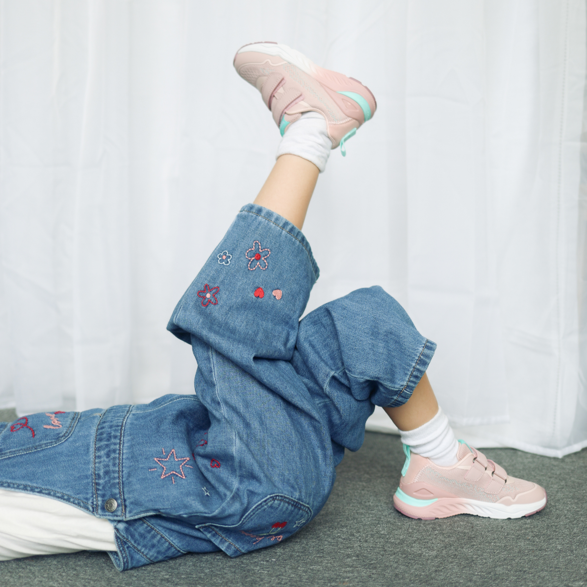 Child lying down wearing pink chunky trainers with mint accents, white socks and embroidered denim jeans.