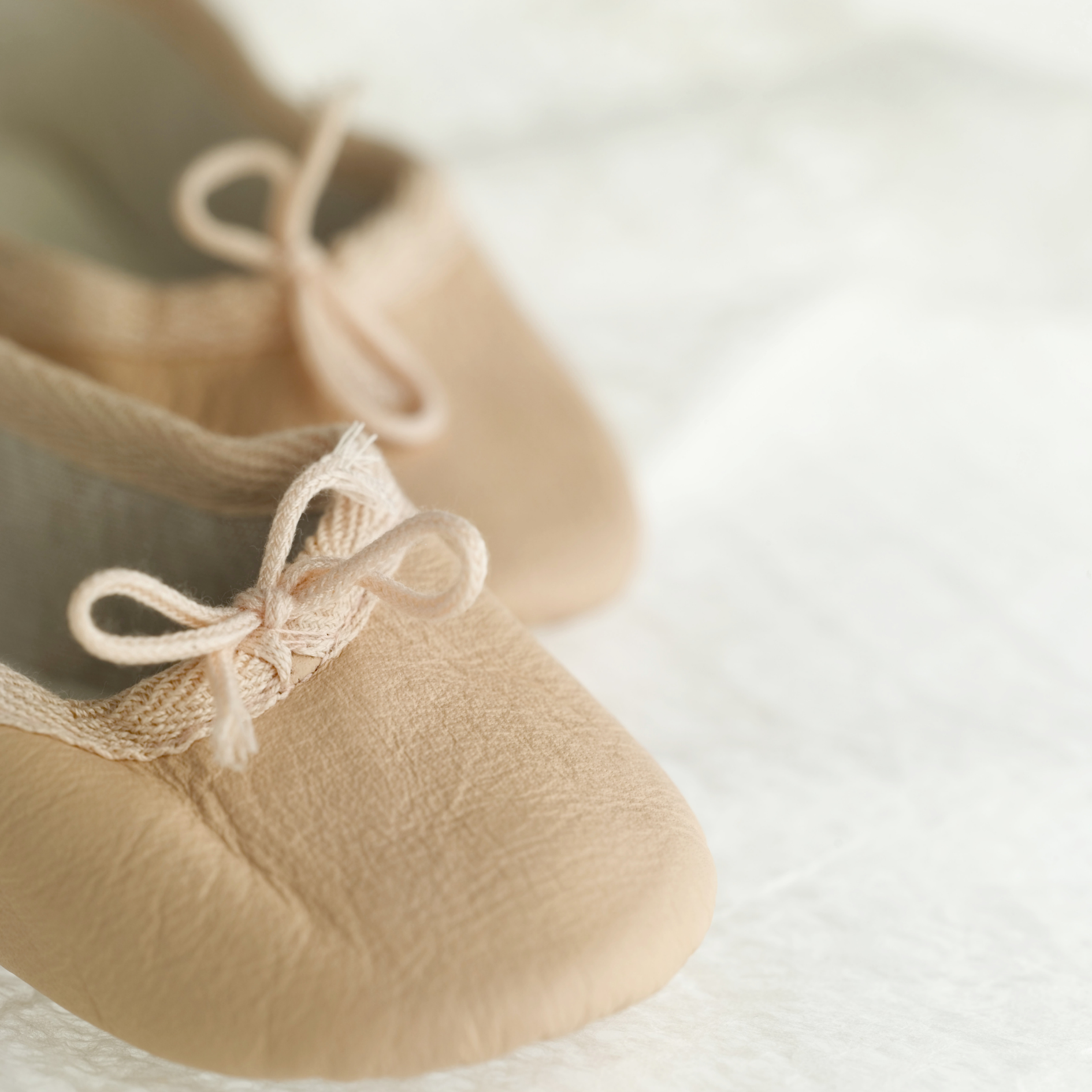 Pair of soft leather baby shoes in blush pink with delicate bow ties, placed on a white fluffy surface.