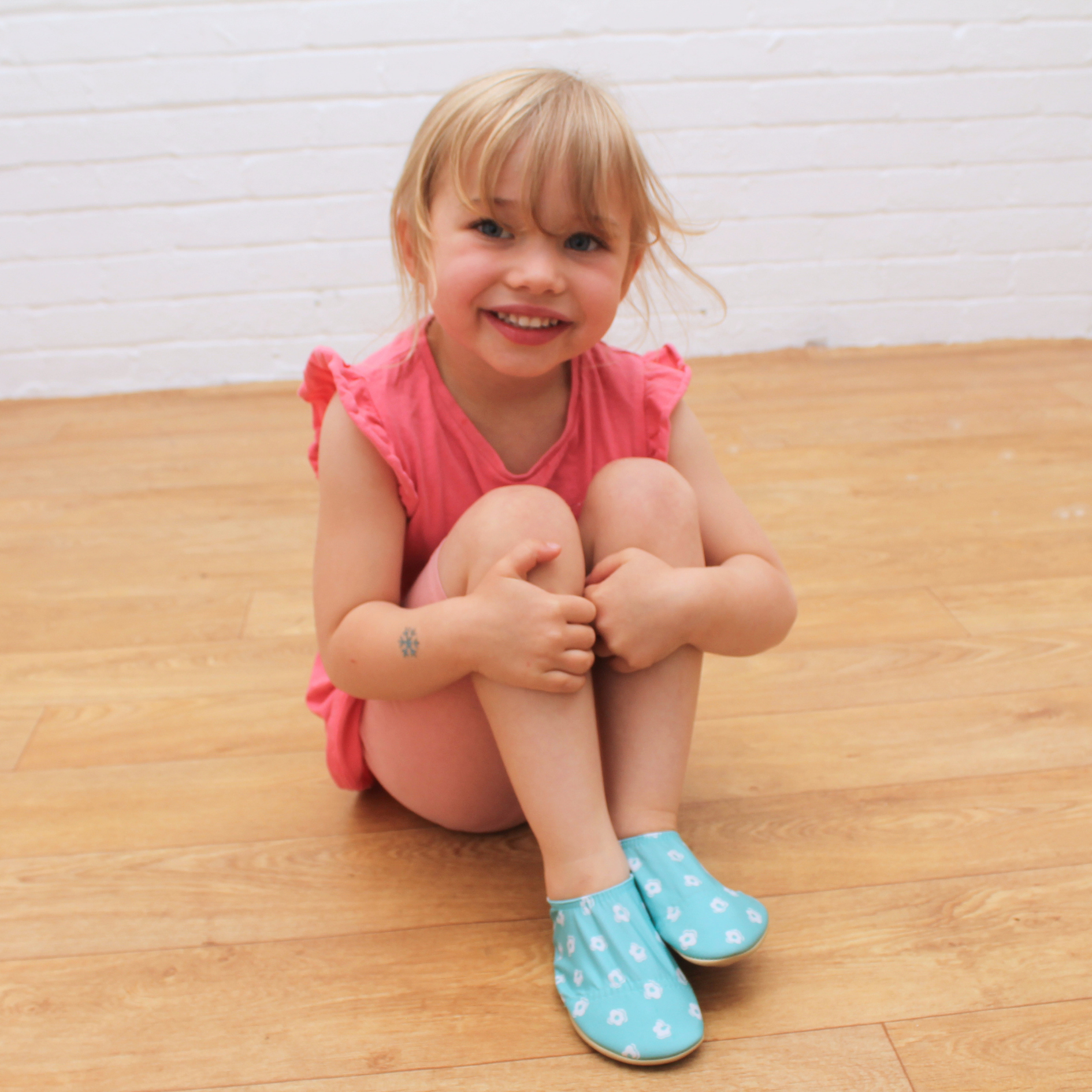 Young girl sitting on floor wearing turquoise floral soft sole shoes with pink dress, smiling at camera.