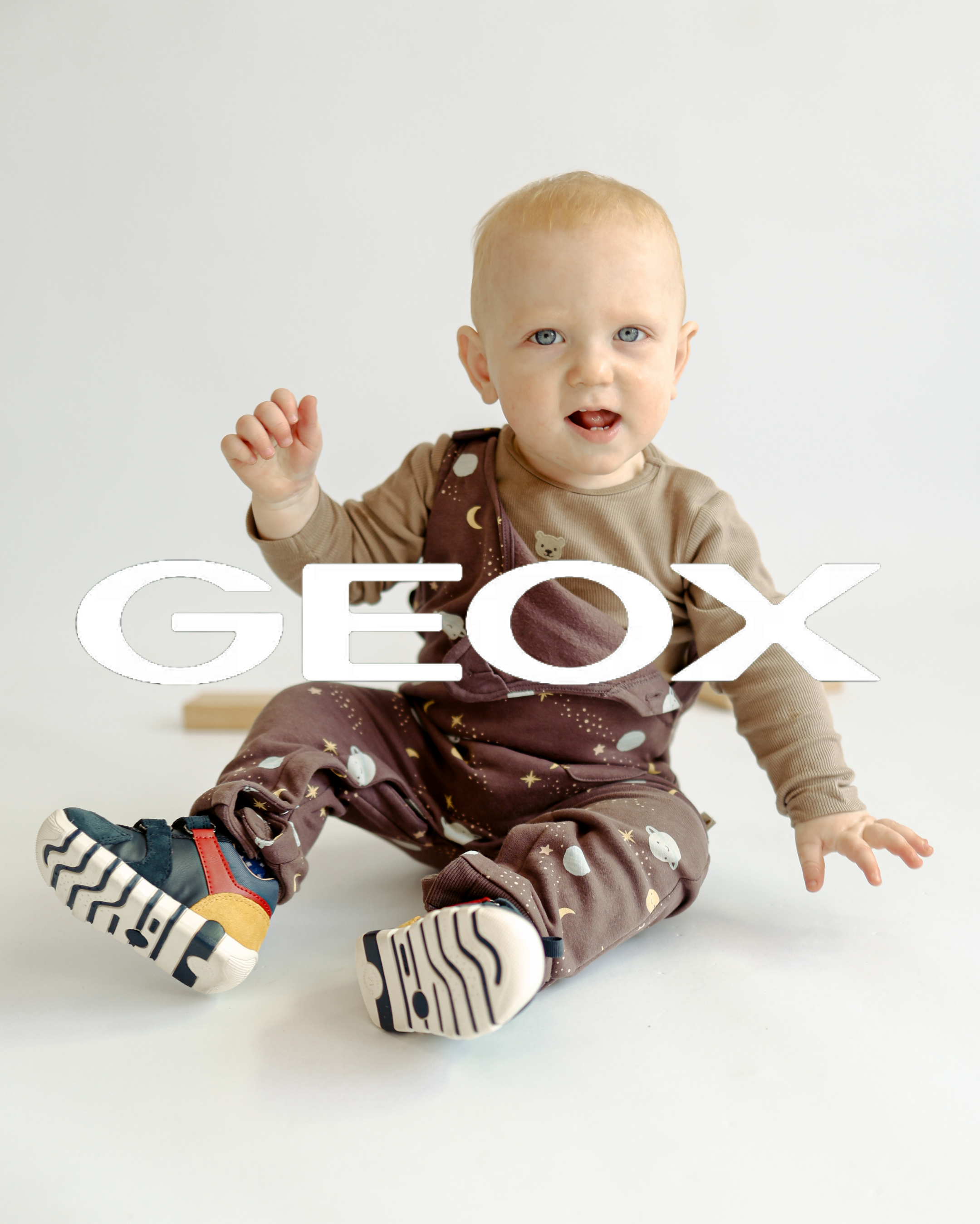 Baby wearing space-print dungarees and colourful Geox first shoes, sitting on white background