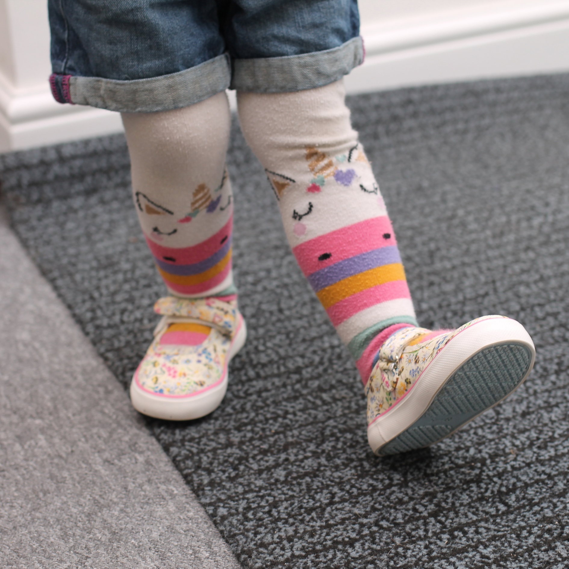 Toddler wearing unicorn knee-high socks and floral canvas shoes with velcro straps, walking on grey carpet.