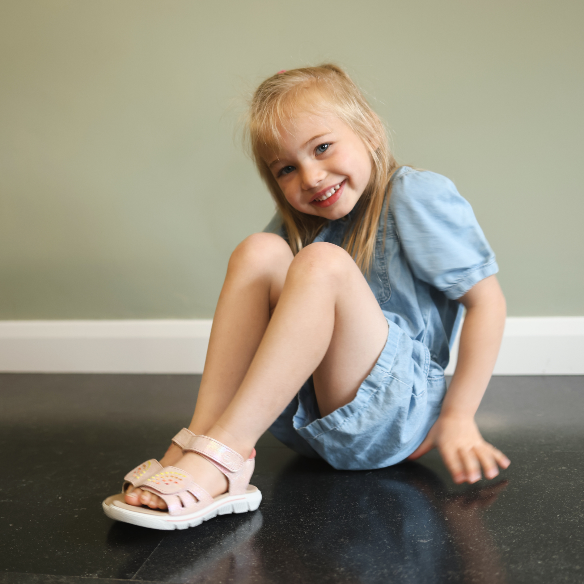 Young girl sitting on floor smiling, wearing pink Skechers sandals with velcro strap and blue denim dress.