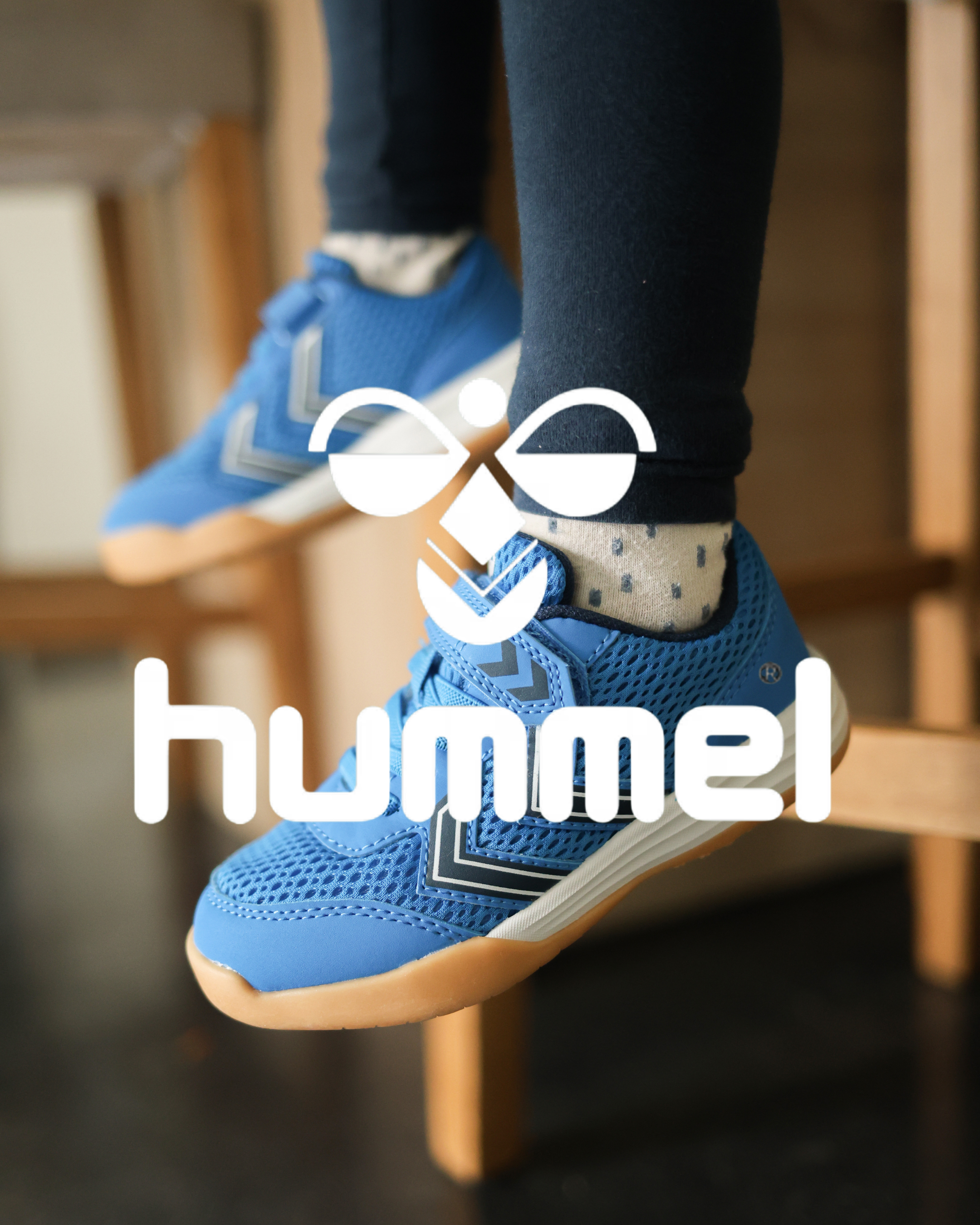 Blue Hummel children's trainers with chevron logo, worn with dark leggings, shown dangling from a wooden bench.