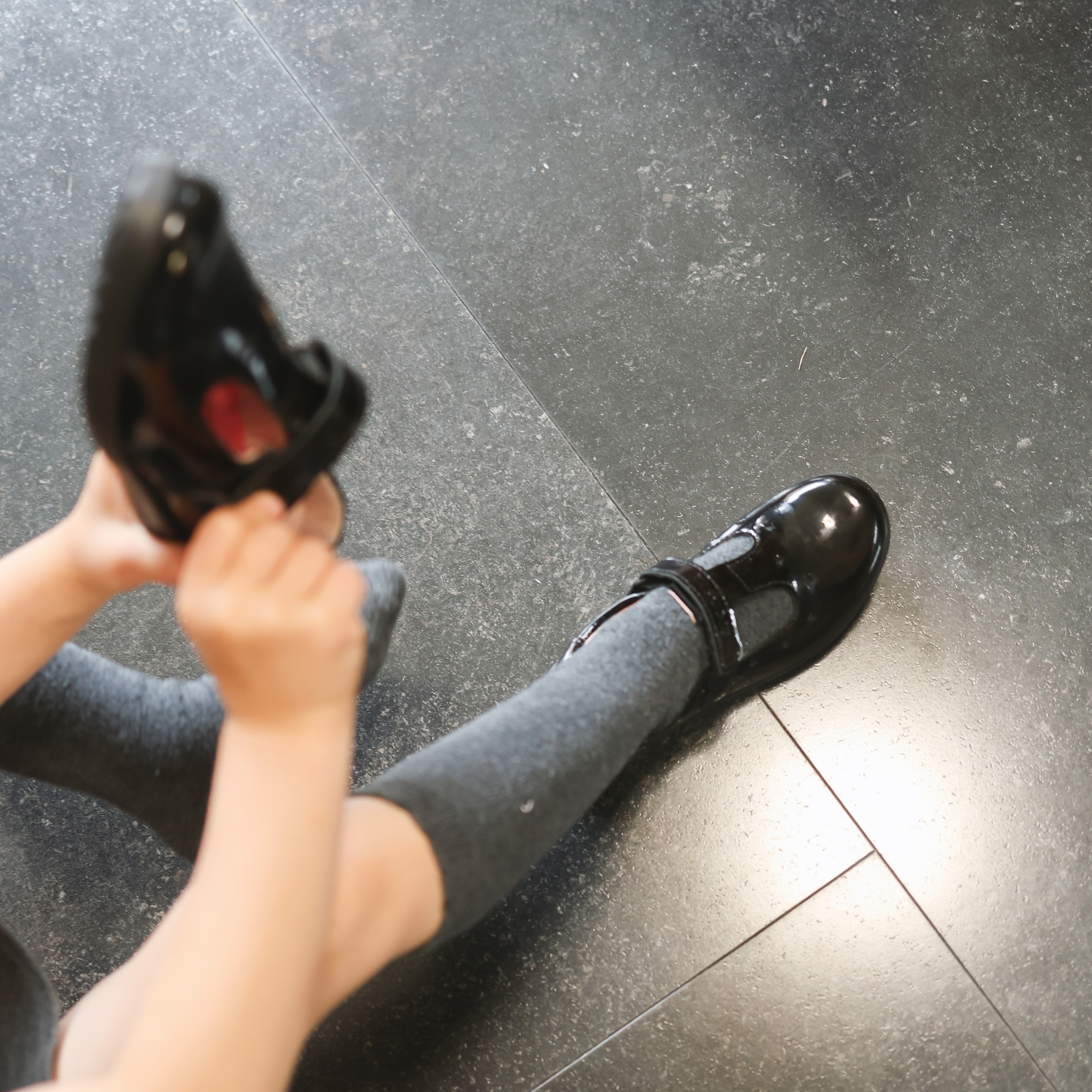 Child putting on black patent leather Mary Jane school shoes with velcro strap, wearing grey socks, seated on tile floor.