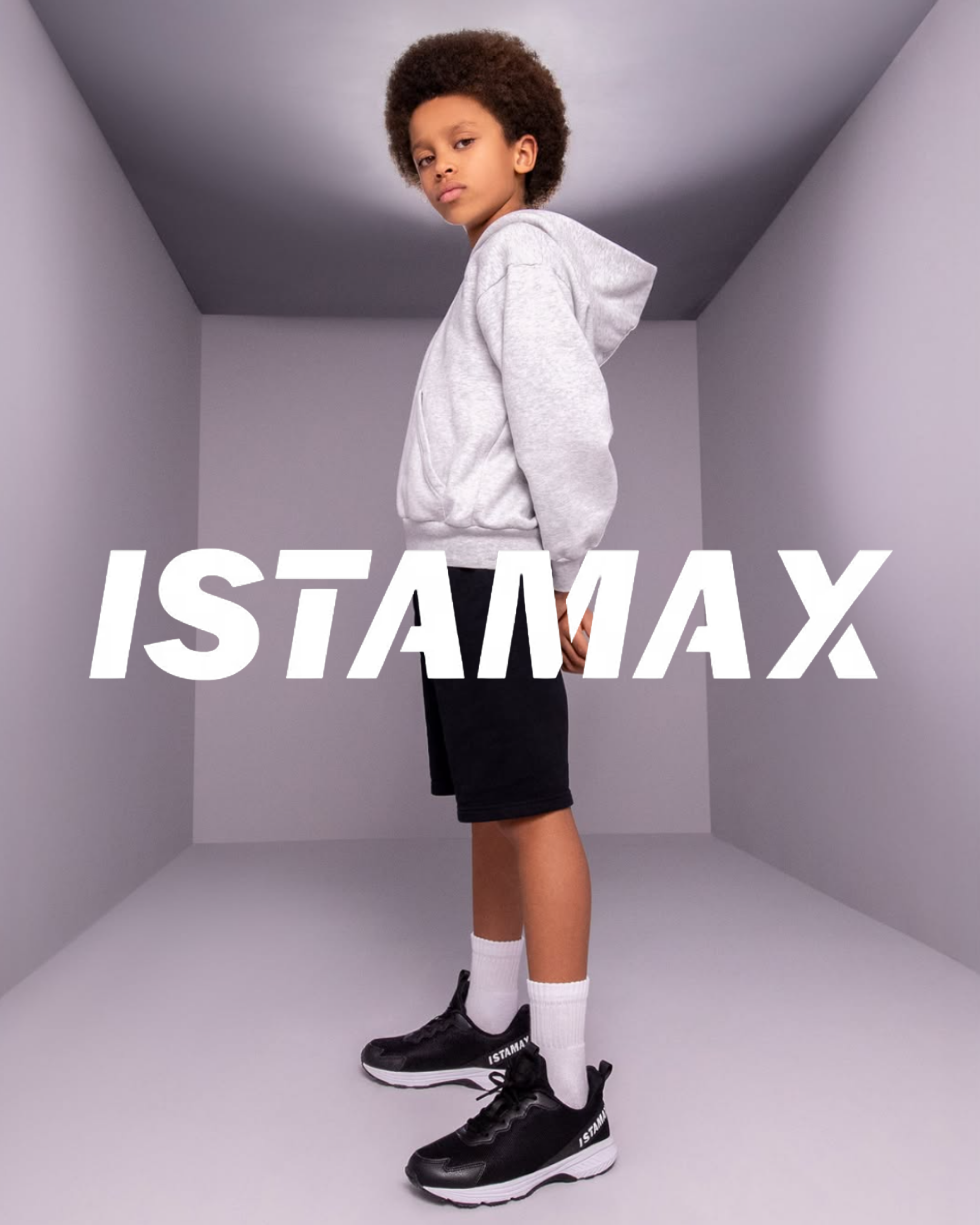 Boy wearing black Istamax trainers with white socks, black shorts and grey hoodie, shown in profile against grey background.