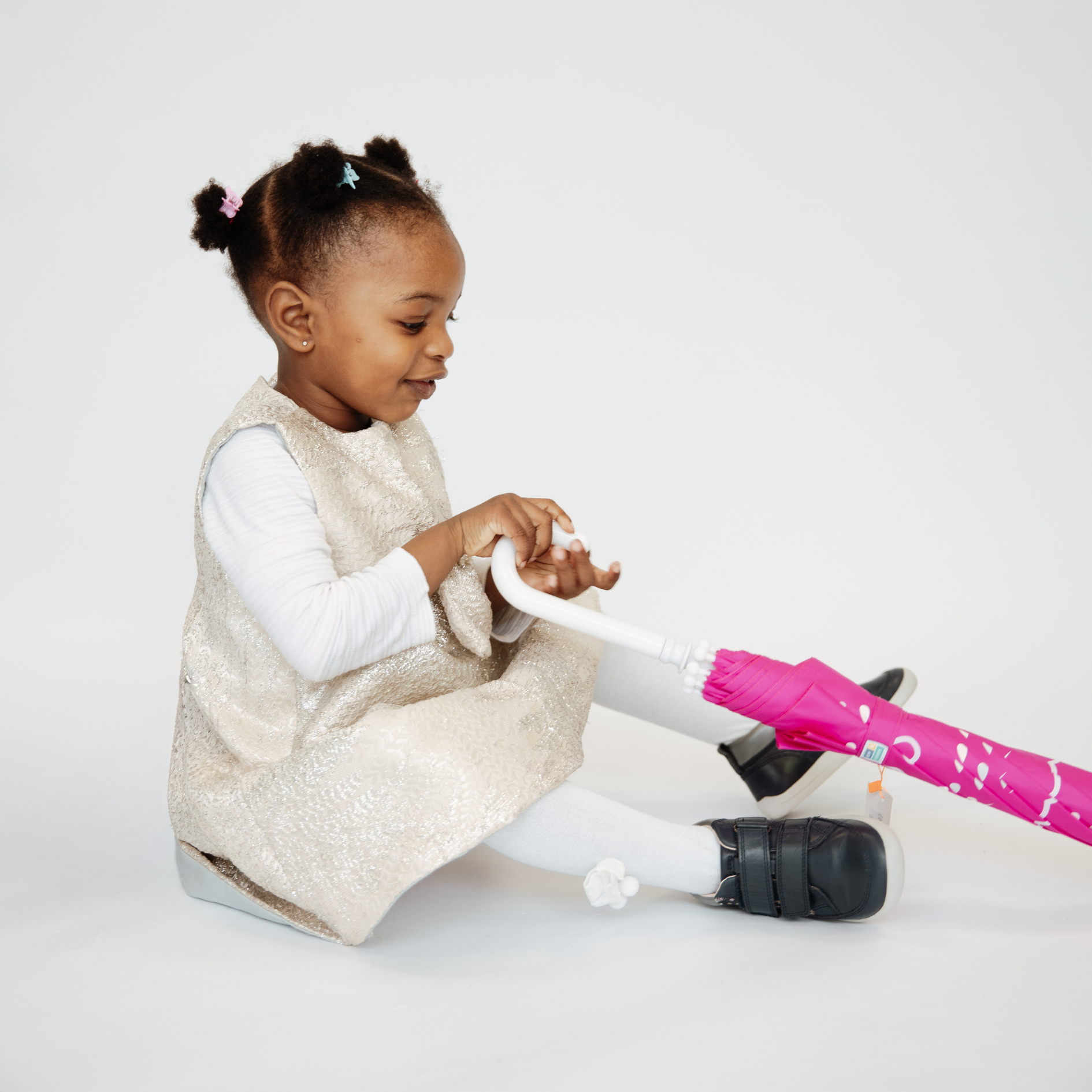 Young girl sitting on floor holding a pink umbrella, wearing orthopaedic shoes with white tights and a silver dress.
