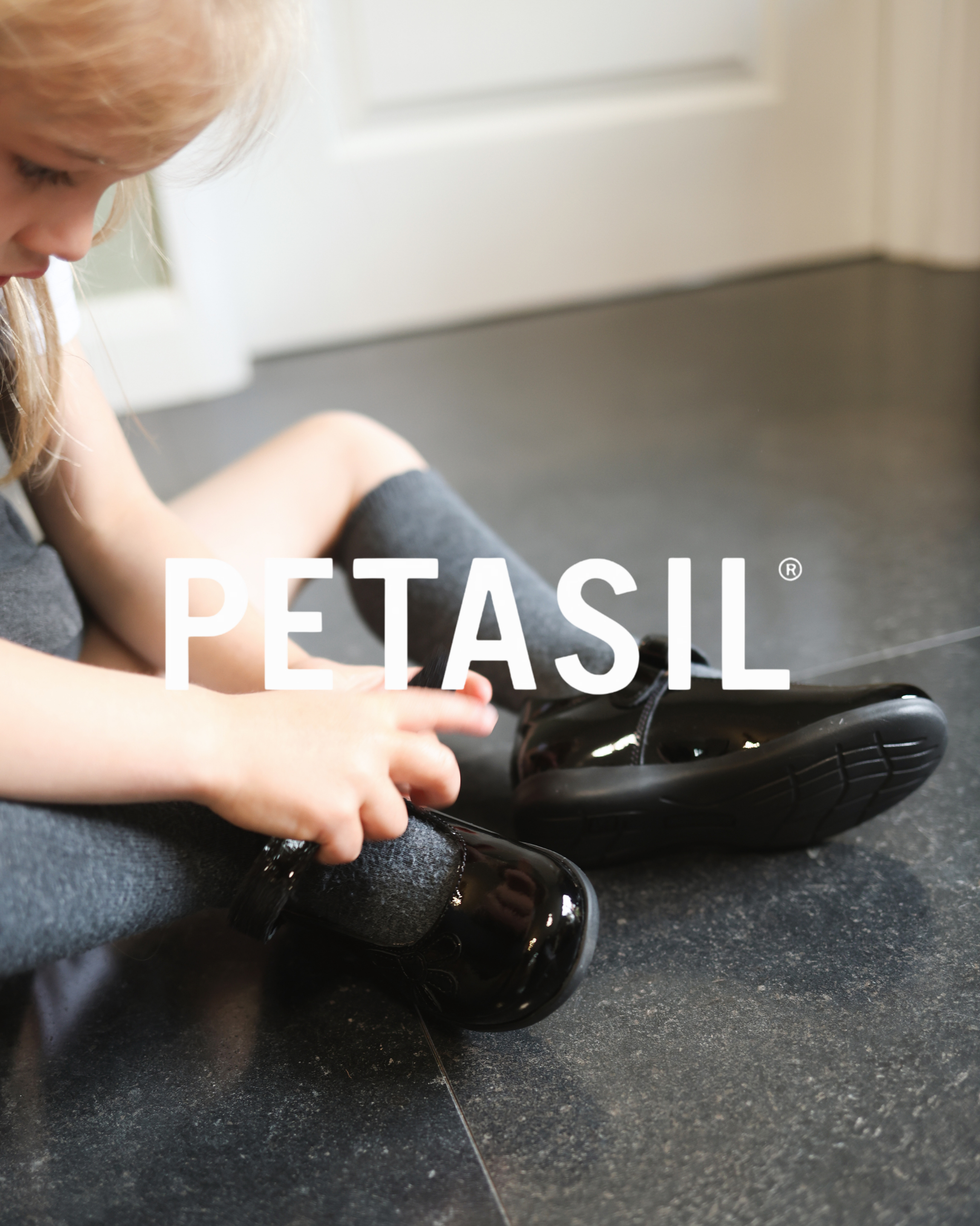 Child putting on shiny black patent leather school shoes by Petasil, sitting on a dark tiled floor