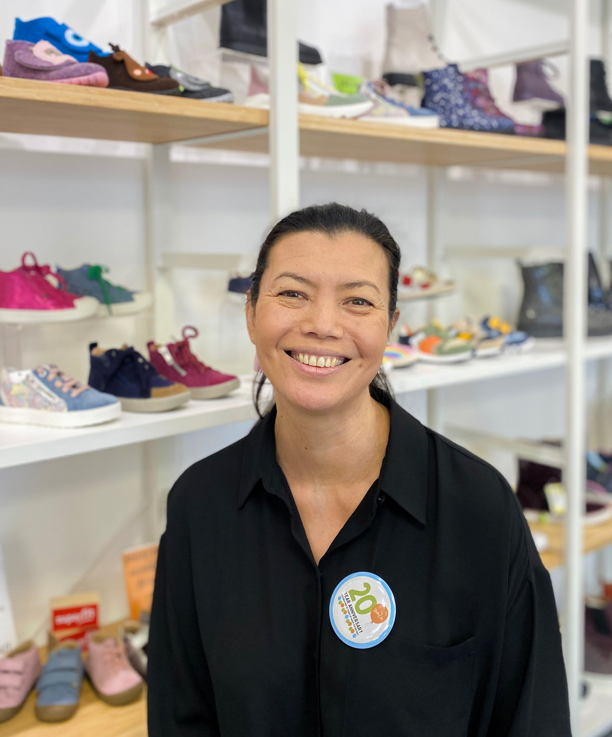 ShuZu staff member smiling in store, wearing black shirt with 20-year anniversary badge, children's shoes on shelves behind