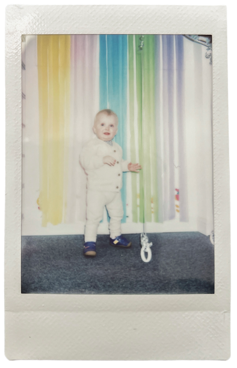 Toddler standing in a white outfit and blue shoes, posing in front of a rainbow-coloured curtain, Instax photo style.