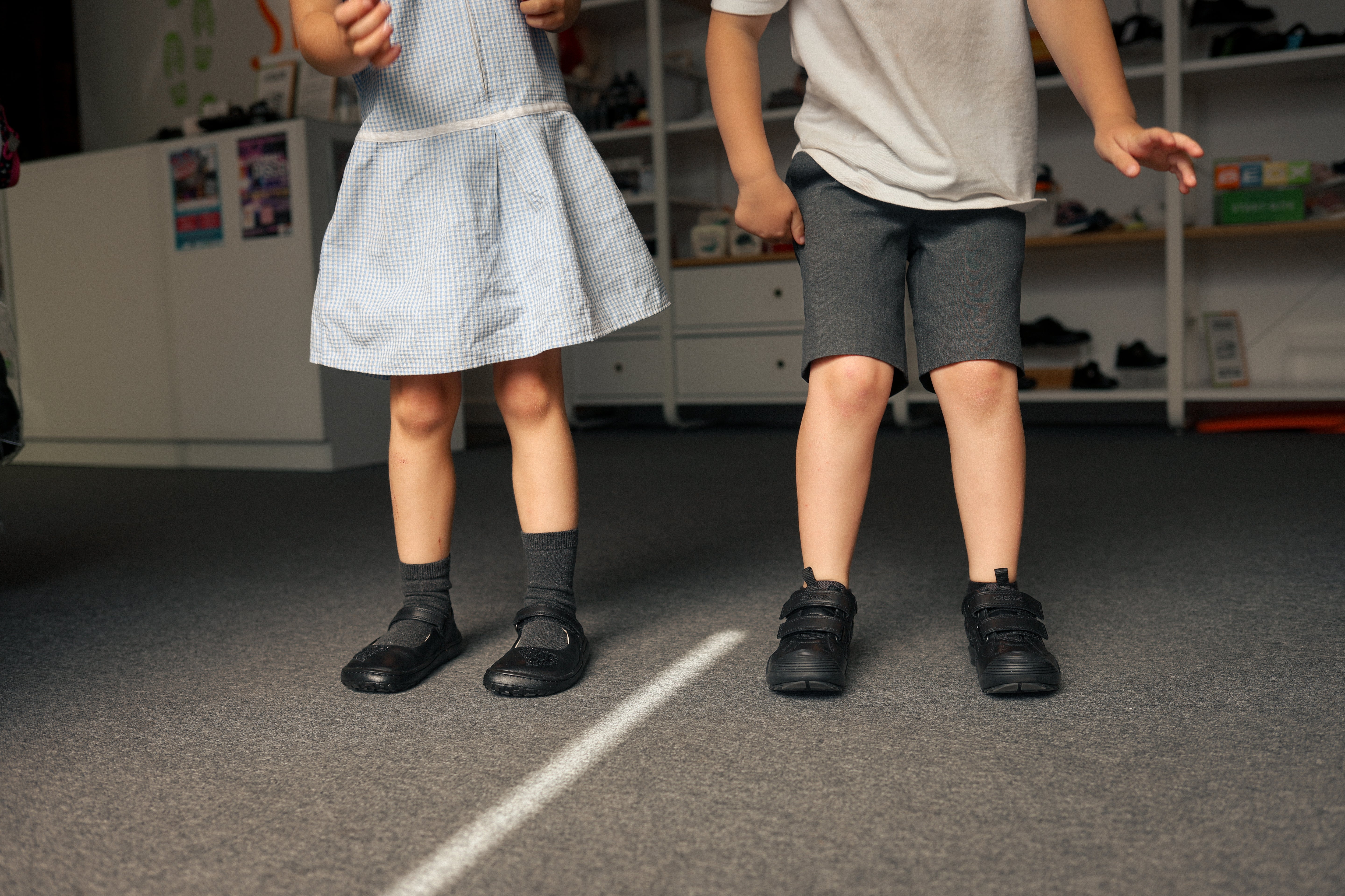 Two young children in school uniforms wearing black velcro school shoes, standing on a carpeted floor.