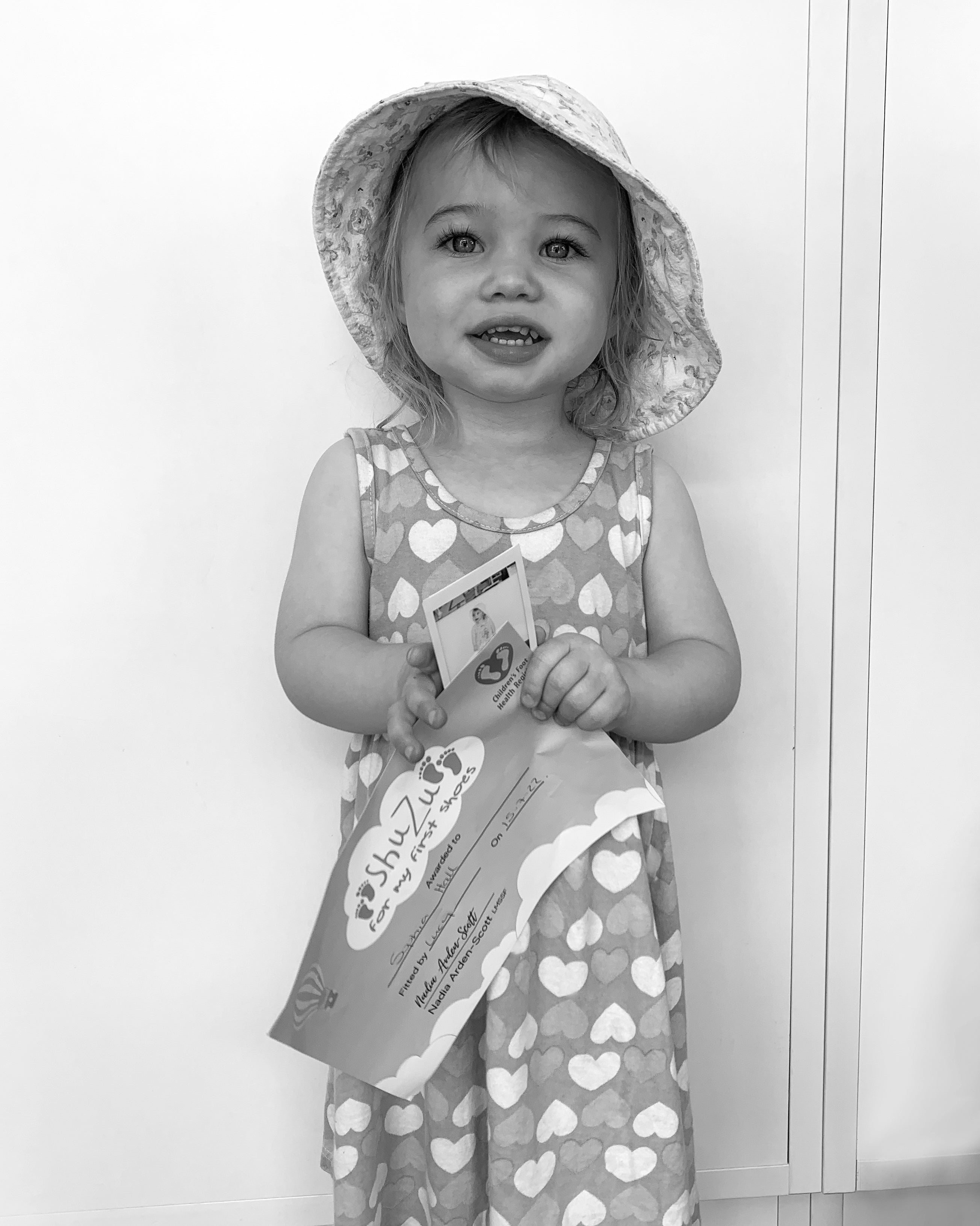 Smiling toddler girl in a sun hat and heart-print dress holding her ShuZu first shoes certificate