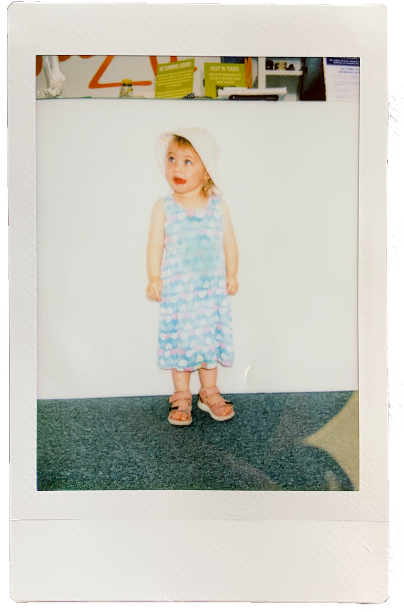 Toddler girl wearing a blue polka dot dress, white sun hat and brown sandals, standing indoors on grey carpet.