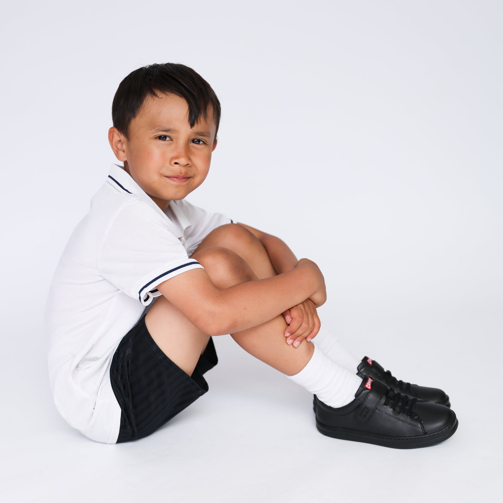 Boy sitting on floor wearing black Camper trainers, white polo shirt, black shorts and white socks.