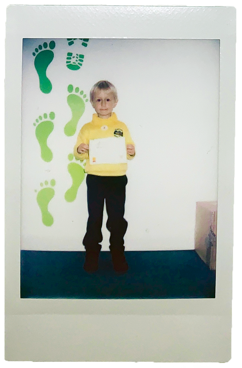 A young child proudly holding a certificate, wearing a yellow school jumper, standing in front of a green footprint wall d...